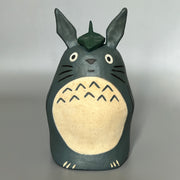 Stylized ceramic figurine of Totoro, standing a smiling with a leaf atop his head.