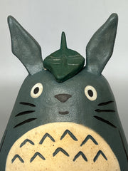 Stylized ceramic figurine of Totoro, standing a smiling with a leaf atop his head.
