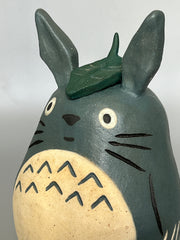 Stylized ceramic figurine of Totoro, standing a smiling with a leaf atop his head.