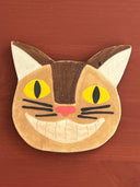 Cat-shaped ceramic coaster with yellow eyes and a pink nose on a wooden surface