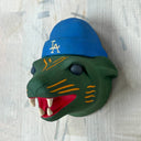 Green tiger head sculpture with a blue cap featuring 'LA' logo on a light wooden background