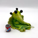 A green, cartoonish slug-like creature figurine with 4 large black eyes and a long snout, posed sitting down next to a miniature red, white, and blue beer can on a white studio background.
