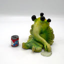 A green, cartoonish slug-like creature figurine with 4 large black eyes and a long snout, posed sitting down next to a miniature red, white, and blue beer can on a white studio background.