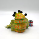 A green, cartoonish slug-like creature figurine with 4 large black eyes and a long snout, posed sitting down next to a miniature red, white, and blue beer can on a white studio background.