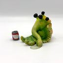 A green, cartoonish slug-like creature figurine with 4 large black eyes and a long snout, posed sitting down next to a miniature red, white, and blue beer can on a white studio background.
