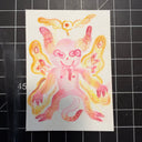 A hand-painted watercolor illustration of a cute, multi-tailed pink monster with yellow and orange wings, shown on a white square card placed on a black gridded cutting mat.