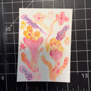 A small watercolor painting of colorful plant shapes on white paper, placed on a black gridded cutting mat with measurement markings visible around it.