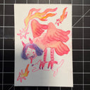 A hand-drawn watercolor and ink illustration on a small white sheet of paper, placed on a black gridded cutting mat. The artwork depicts a whimsical scene with a pink bird-like figure, flames, stars, and a purple-haired horned character.