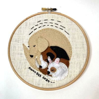 Embroidery in a hoop of 3 dogs, nestled into one another from smallest to largest. Dogs are different shades of brown, black and white. 