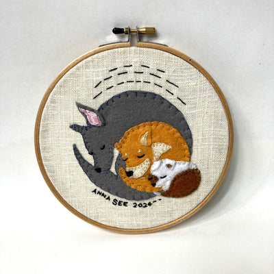Embroidery in a hoop of 3 dogs, nestled into one another from smallest to largest. Dogs are different shades of brown, gray and white.