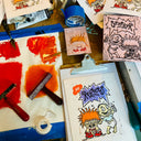 A tabletop display of hand-printed cartoon art and printing tools, including a clipboard with a printed illustration, wood blocks, a roller, ink swatches in red and orange, blue tape, and sketch papers.