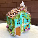 Paper sculpture of a 3D two story house, with shingle roofing, teal colored window shutters and ivy growing along the side. Cats crawl on the flower window boxes and tissues can be pulled from the chimney. 