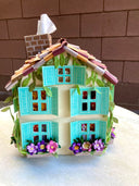 Paper sculpture of a 3D two story house, with shingle roofing, teal colored window shutters and ivy growing along the side. Cats crawl on the flower window boxes and tissues can be pulled from the chimney. 