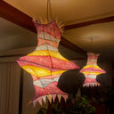 A hanging paper lantern with a geometric, layered paper or fabric design in pink, yellow, and white, suspended in a dimly lit room with wooden ceiling beams and plants below.