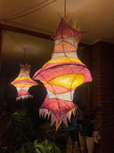 A hanging decorative lantern with a vaguely fish-like silhouette, glowing from within. The exterior is made of layered translucent paper or fabric in pink, red, yellow, and white, with pointed triangular edges.