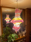 A hanging decorative lantern with a vaguely fish-like silhouette, glowing from within. The exterior is made of layered translucent paper or fabric in pink, red, yellow, and white, with pointed triangular edges.