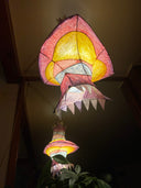 A hanging decorative lantern with a vaguely fish-like silhouette, glowing from within. The exterior is made of layered translucent paper or fabric in pink, red, yellow, and white, with pointed triangular edges.