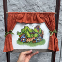 Framed illustration of a green fish monster laying atop of houses and playing the banjo. Framed illustration has a red curtain around it, as though revealing the artwork as a stage.