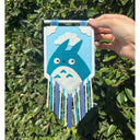 Hand holding a decorative banner with a blue Totoro character against a green leafy background. Banner has strips of fabric.