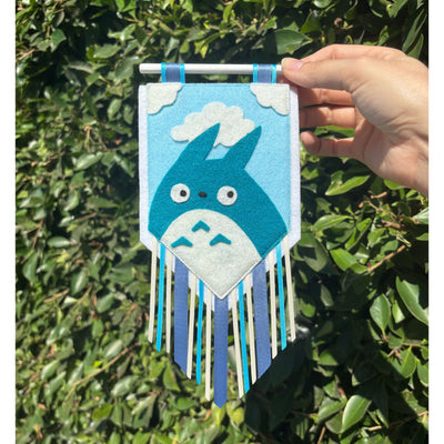 Hand holding a decorative banner with a blue Totoro character against a green leafy background. Banner has strips of fabric.
