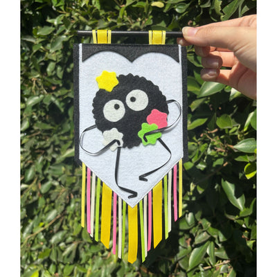 Felt banner with a decoration of a soot sprite, carrying several colorful stars. Fabric strips hang from the banner.