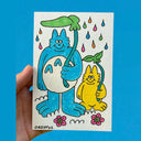 Marker drawing on paper of a blue large cat creature with big feet and hands, holding up a leaf umbrella over its head. Next to it is a smaller yellow cat creature, holding up a similar umbrella.