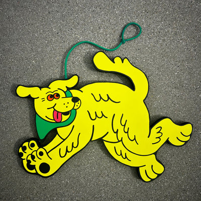 Die cut wooden painting of a yellow dog with a green bandana and leash, running and looking excitedly behind it.