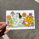 A hand holds a white artwork featuring a colorful cartoon illustration of a light green dog and a yellow bird holding hands and surrounded by orange and pink flowers, with an asphalt street background.