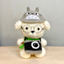 Fluffy plush sculpture of a white dog with black beaded eyes and wire framed glasses. It wears a green scarf and has a camera around its neck and a Totoro hat.