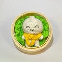 Crocheted white character with a yellow scarf inside a bamboo steamer on a light gray background