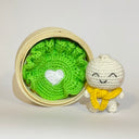 Crocheted white character with a yellow scarf next to a crocheted leaf of lettuce in a bamboo steamer basket.