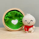 Crochet sculpture of a bao bun with a cute, smiling face and a tiny body. It wears a red scarf and sits next to a crocheted bed of lettuce with a white heart center, inside of a real steamer basket.