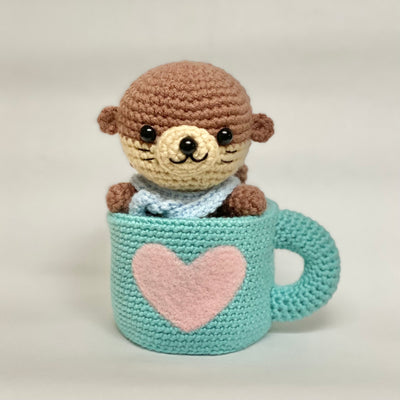 Crocheted otter plush inside a teal mug with a pink heart on a white background
