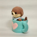 Crocheted otter plush inside a teal mug with a pink heart on a white background