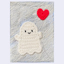 Texture white paper with a yarn sculpture on top of a ghost holding a heart shaped red balloon.