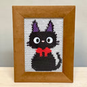 Framed cross-stitched picture of a black cat with purple ears and a red bow tie on a white background.