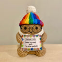 Crocheted sculpture of a small brown potato with short arms and legs. It has glasses, wears a rainbow colored beanie and holds a sign that reads "positive potato"