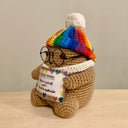 Crocheted sculpture of a small brown potato with short arms and legs. It has glasses, wears a rainbow colored beanie and holds a sign that reads "positive potato"