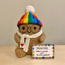 Crocheted sculpture of a small brown potato with short arms and legs. It has glasses, wears a rainbow colored beanie and holds a sign that reads "positive potato"