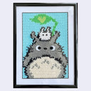 Framed cross stitch artwork of Totoro with a tiny white Tototo atop of it with a green leaf.
