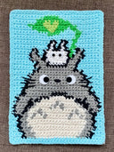 Fabric cross stitch artwork of Totoro with a tiny white Tototo atop of it with a green leaf.