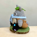 Crochet knitting sculpture of Totoro, wearing a blue scarf and a green leaf atop his head next to 2 soot sprites with stars attached. They are atop of a wood slice base with a green crochet rug atop.