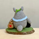 Crochet knitting sculpture of Totoro, wearing a blue scarf and a green leaf atop his head next to 2 soot sprites with stars attached. They are atop of a wood slice base with a green crochet rug atop.