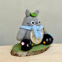 Crochet knitting sculpture of Totoro, wearing a blue scarf and a green leaf atop his head next to 2 soot sprites with stars attached. They are atop of a wood slice base with a green crochet rug atop.