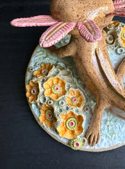 Ceramic sculpture of a cute axolotl, brown in color with pink color accents. It looks up at the viewer and is on a base of coral.