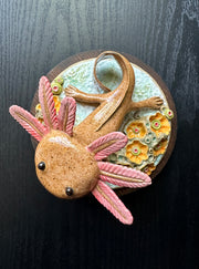 Ceramic sculpture of a cute axolotl, brown in color with pink color accents. It looks up at the viewer and is on a base of coral.