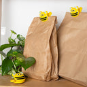 Three yellow bee-shaped clips on brown paper bags with a plant in the background.