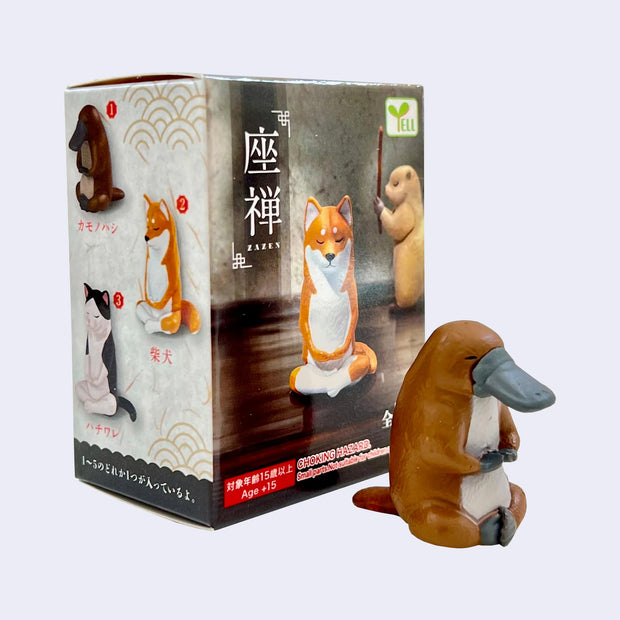 Small plastic platypus, brown with a white stomach and gray bill and paws. It sits in a meditative pose next to its product packaging.