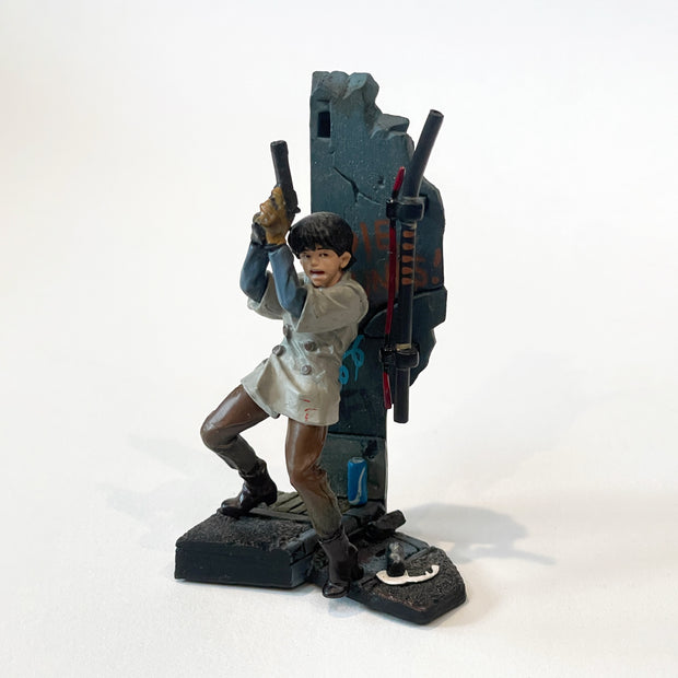 Vinyl figure of a character from Akira holding a gun and standing back to a concrete wall. He has an open mouth expression and pipes come out of the platform, the sign overhead is missing so an empty slot remains.