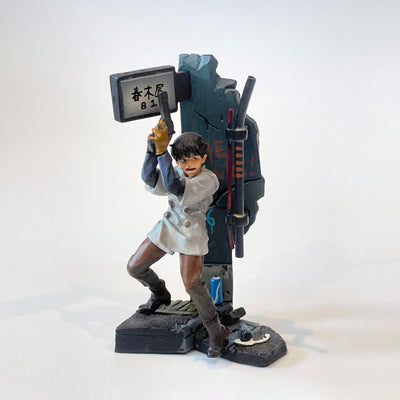 Vinyl figure of a character from Akira holding a gun and standing back to a concrete wall. He has an open mouth expression and pipes come out of the platform, with a light up sign overhead.